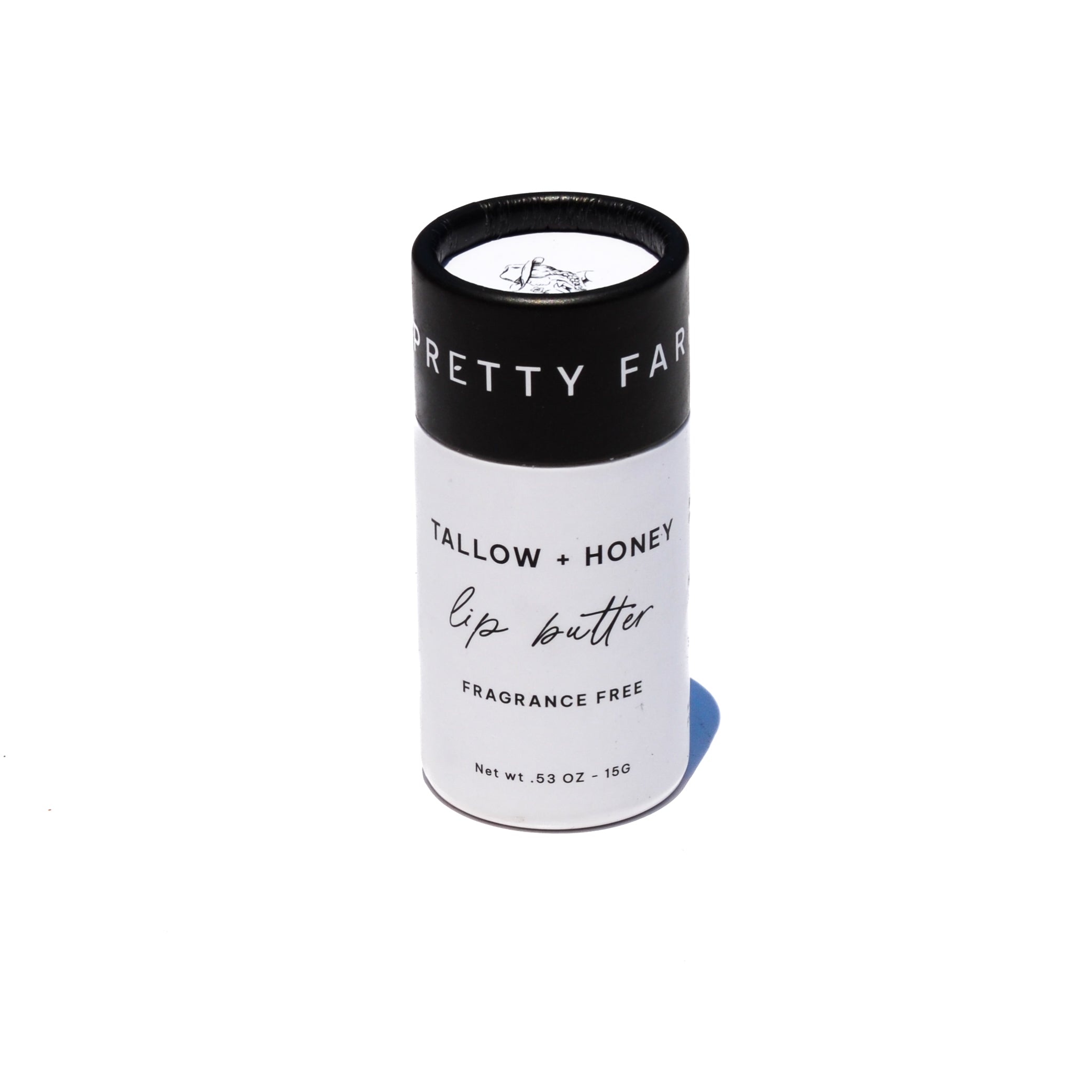 A white lip butter stick labeled 'TALLOW + HONEY' stands on a rustic wooden surface beside a black cap and honeycomb, with a burlap cloth and blurred outdoor field background. grass fed lip balm pretty farm girl handmade skin organic plastic free dry cracked lips repair comfrey