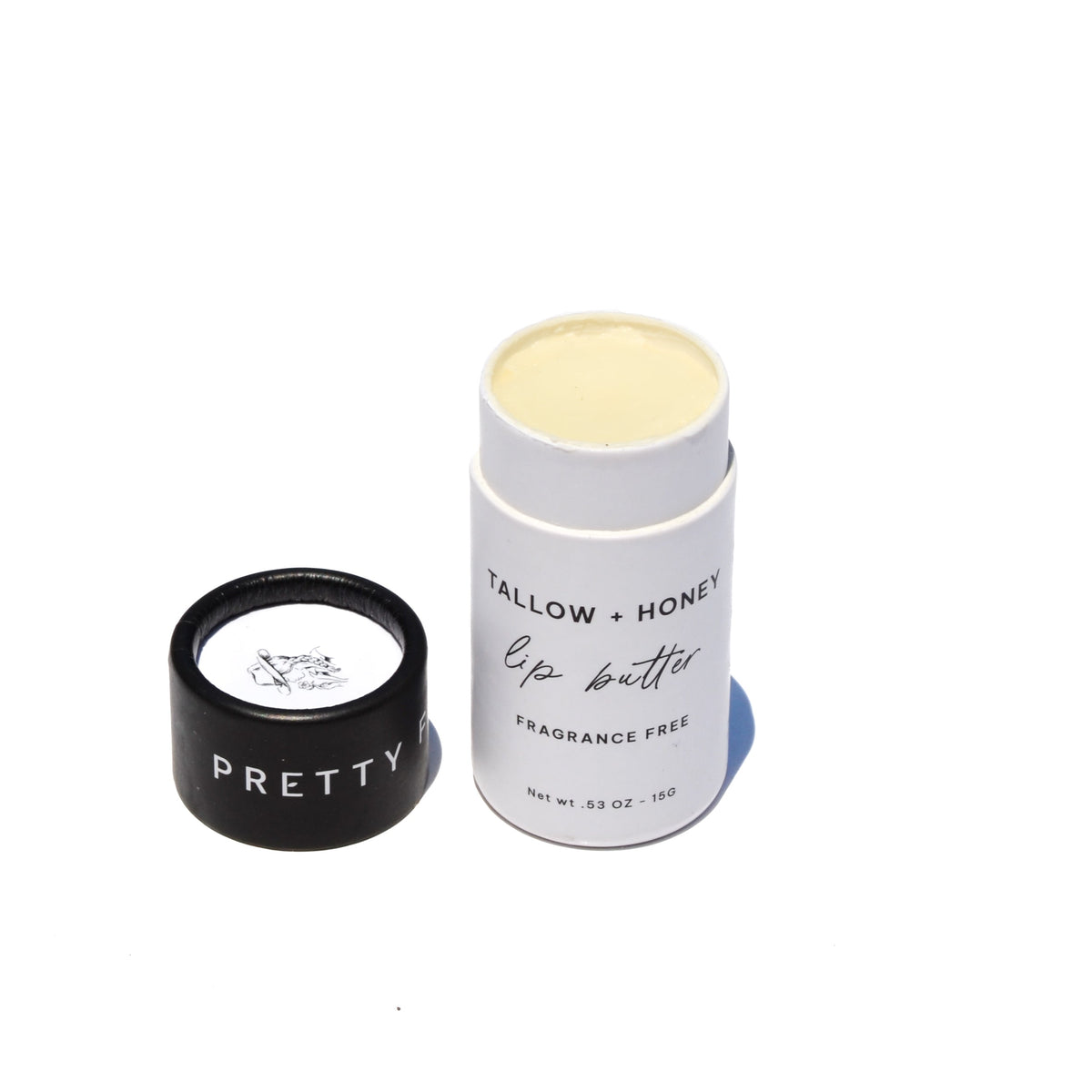 A white lip butter stick labeled 'TALLOW + HONEY' stands on a rustic wooden surface beside a black cap and honeycomb, with a burlap cloth and blurred outdoor field background. grass fed lip balm pretty farm girl handmade skin organic plastic free dry cracked lips repair comfrey