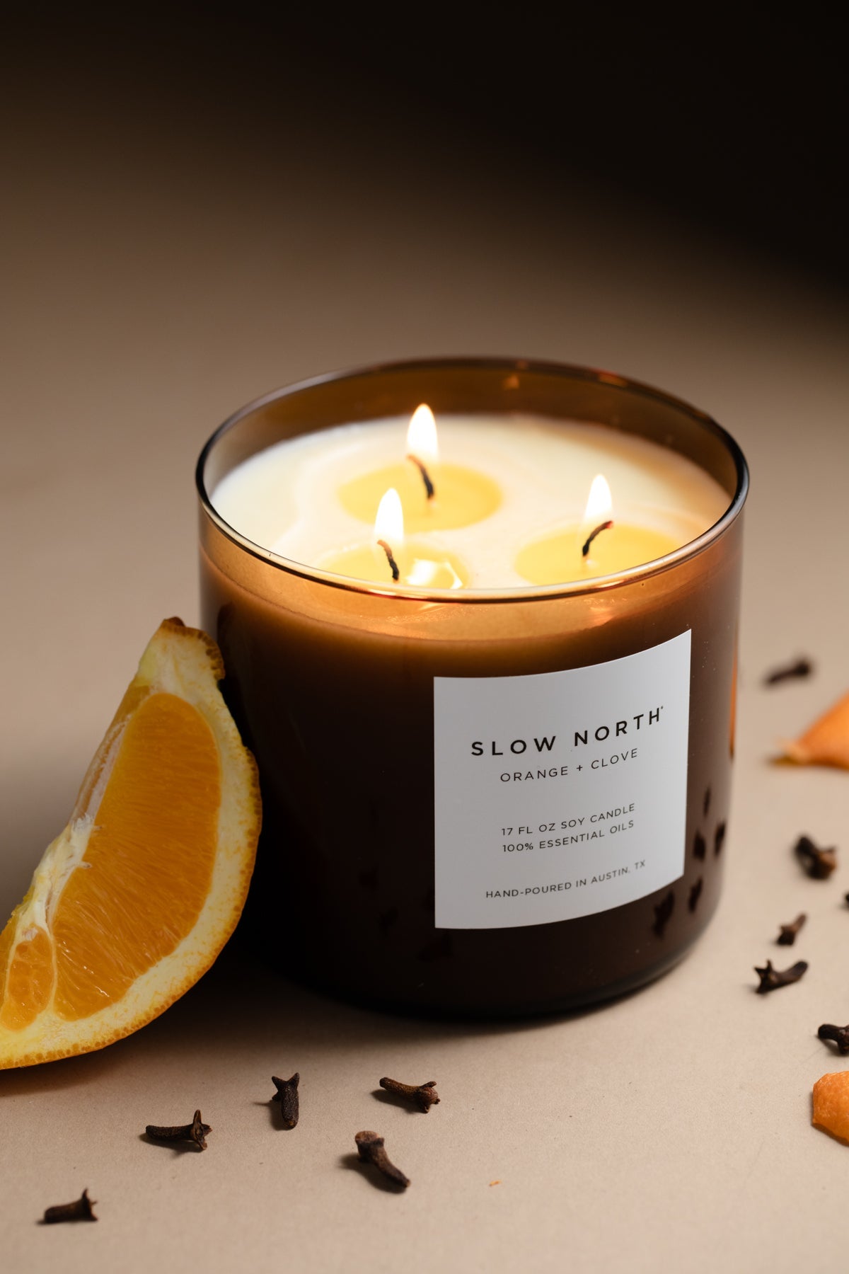 Lit candle with a 'Slow North' label, surrounded by an orange wedge and cloves on a neutral background.Lit candle in a glass jar with 'Slow North' label on a marble surface. SSF Organics Natural beeswax candles with essential oils holiday scents organic holistic relaxing