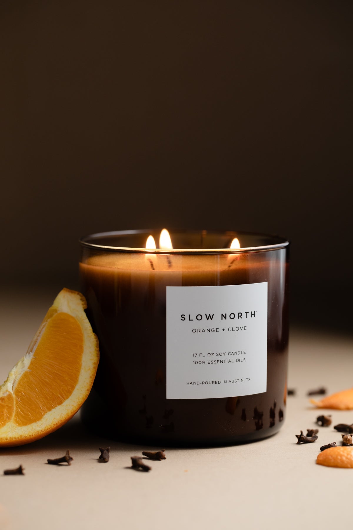 Candle with 'Slow North' label next to an clove and an orange slice on a dark background. Lit candle in a glass jar with 'Slow North' label on a marble surface. SSF Organics Natural beeswax candles with essential oils holiday scents organic holistic relaxing
