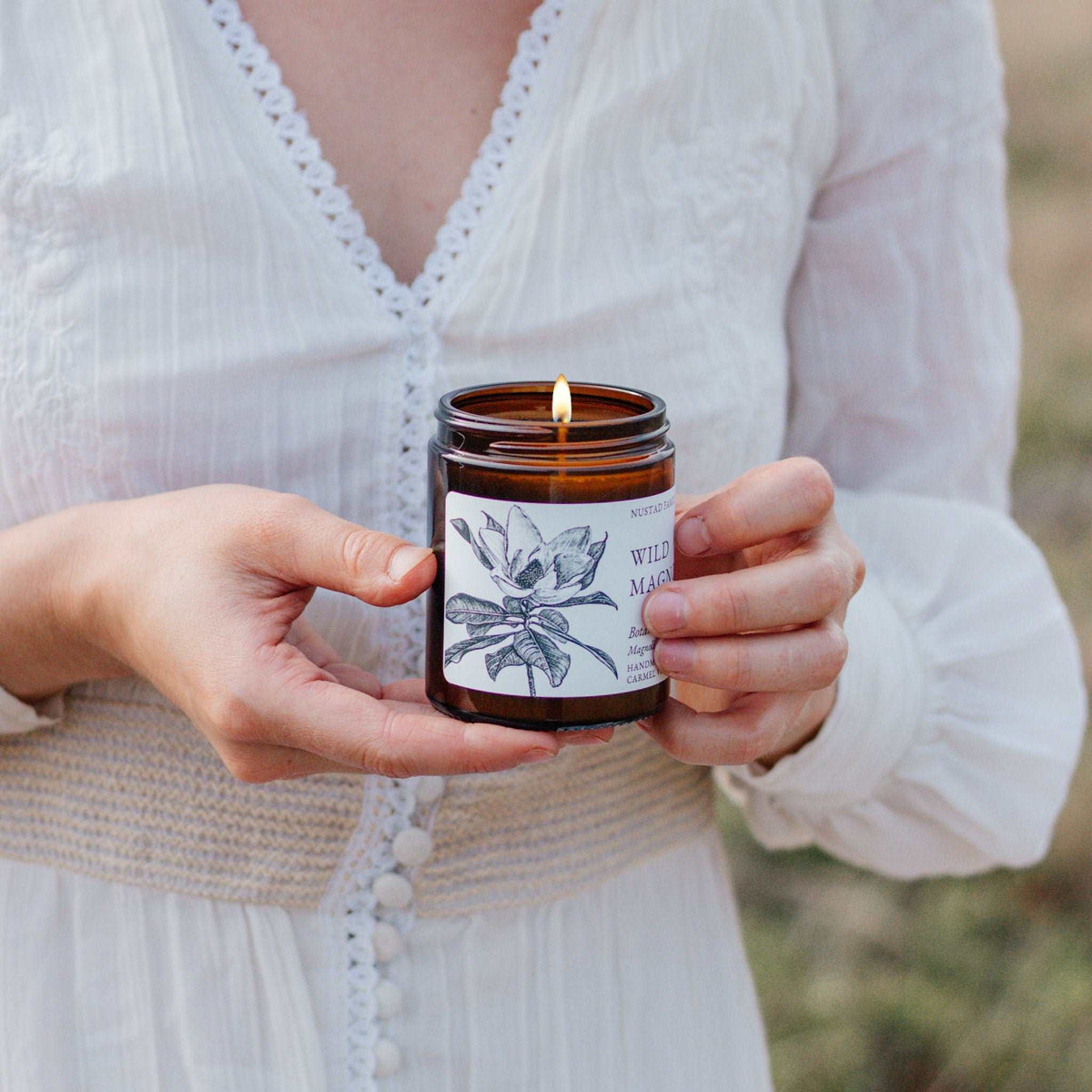 Close-up of hands holding a lit Wild Magnolia candle in an amber glass jar with a floral label.