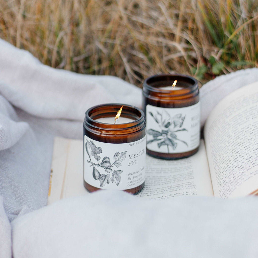 Two lit botanical candles in amber glass jars resting on an open book outdoors.