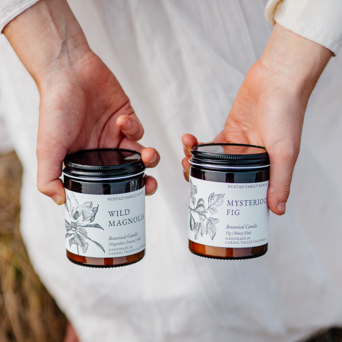 Hands holding Wild Magnolia and Mysterious Fig botanical candles in amber jars with black lids.