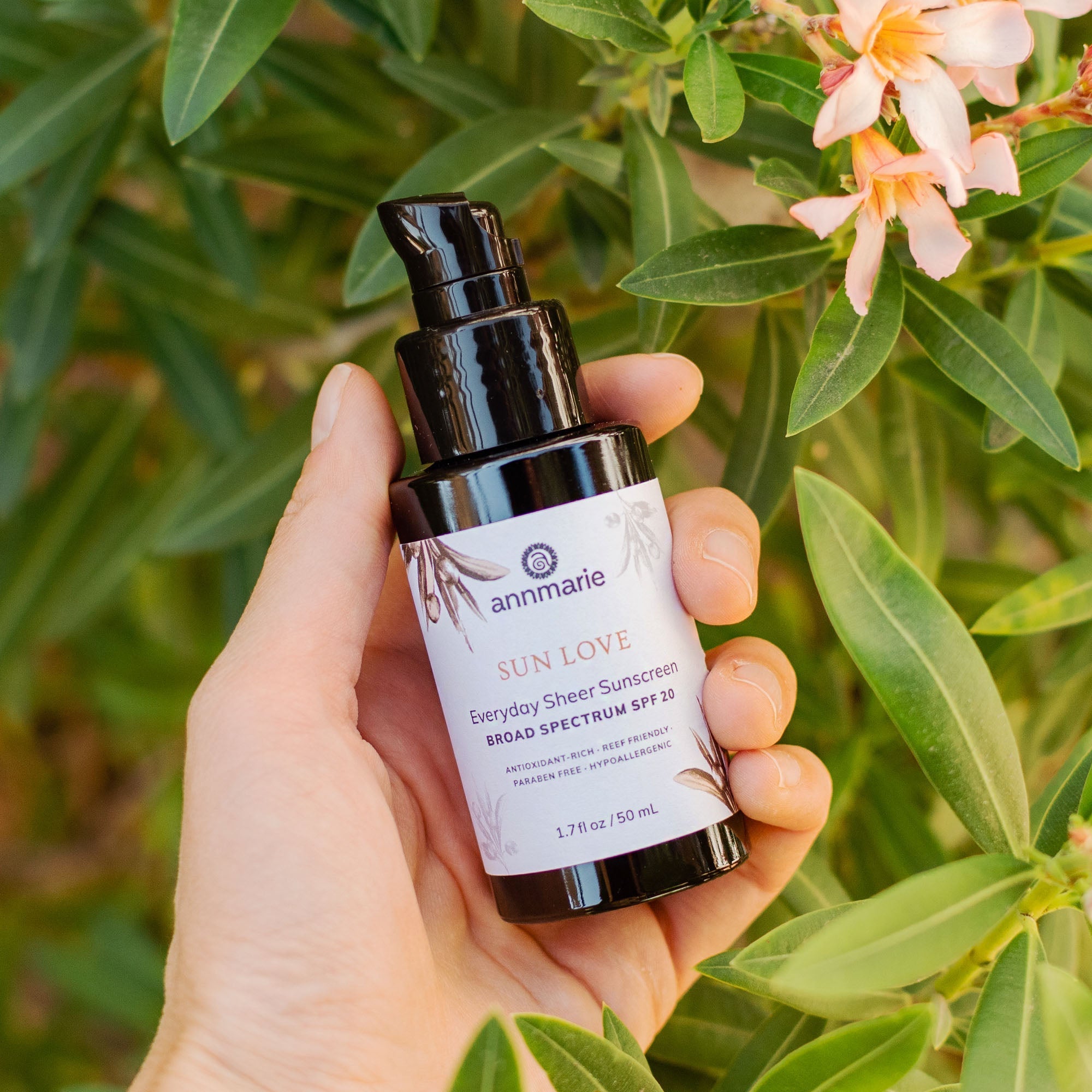 Hand holding a bottle of 'Sun Love' sunscreen with green foliage and flowers in the background. Annmarie Sun Love sunscreen bottle with ingredients listed on a white background. Hand holding a bottle of 'Sun Love' sunscreen with green foliage in the background. natural skin face sunscreen. comfrey and arnica tallow