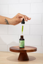 Hand using a dropper to extract liquid from a bottle labeled 'Neem Oil' on a wooden stand with a white tiled wall background.
