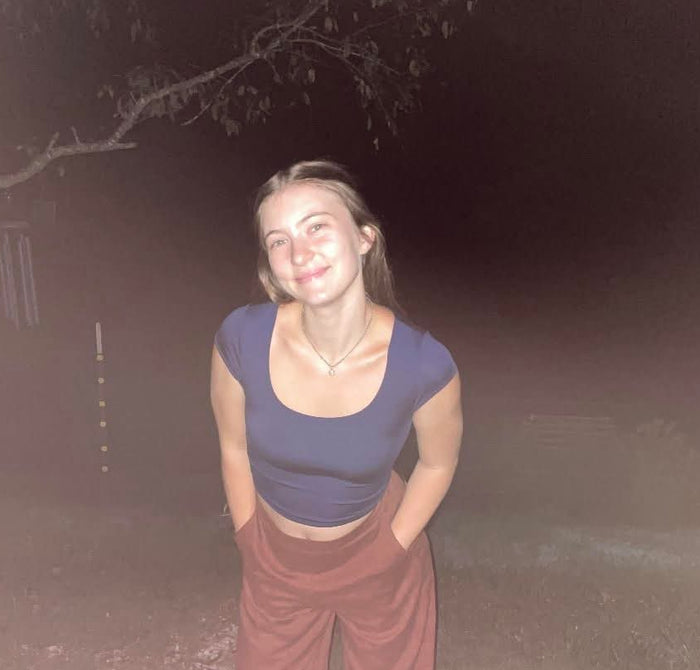 A young woman posing outdoors at night wearing a dark purple scoop-neck crop top and rust-colored wide-leg pants.