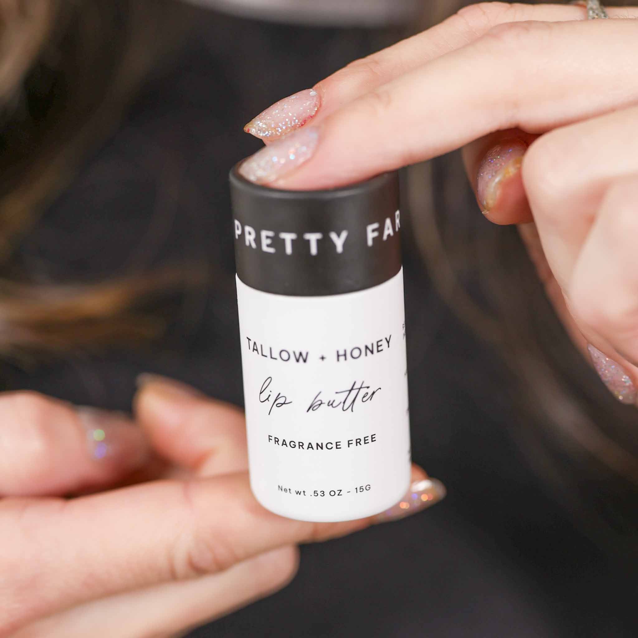 A white lip butter stick labeled 'TALLOW + HONEY' stands on a rustic wooden surface beside a black cap and honeycomb, with a burlap cloth and blurred outdoor field background. grass fed lip balm pretty farm girl handmade skin organic plastic free dry cracked lips repair comfrey
