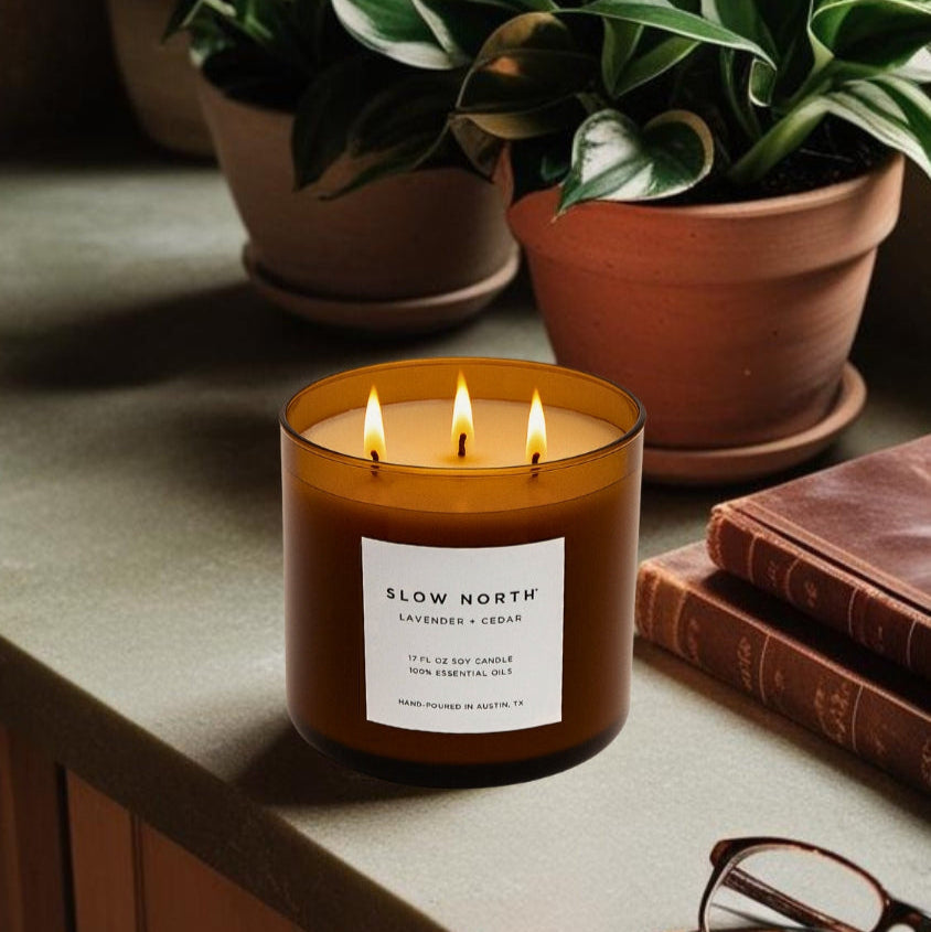 Candle with 'Slow North' label on a surface with potted plants and books in the background. Transform your space into a serene retreat with our new&nbsp;Signature 3-Wick Candle in Lavender + Cedar—a calming, grounded blend designed to fill larger spaces with soft, natural fragrance and a warm, even glow. Topped with a black metal lid.

Hand-poured in Austin, Texas, this candle is crafted with 100% soy wax and pure essential oils, offering a clean, non-toxic burn you can feel good about.