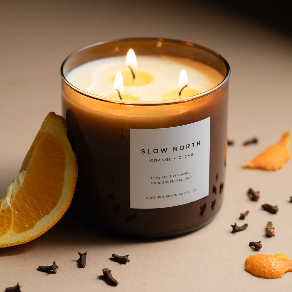 Scented candle labeled 'Slow North' with orange and clove around it on a neutral background. Lit candle in a glass jar with 'Slow North' label on a marble surface. SSF Organics Natural beeswax candles with essential oils holiday scents organic holistic relaxing 