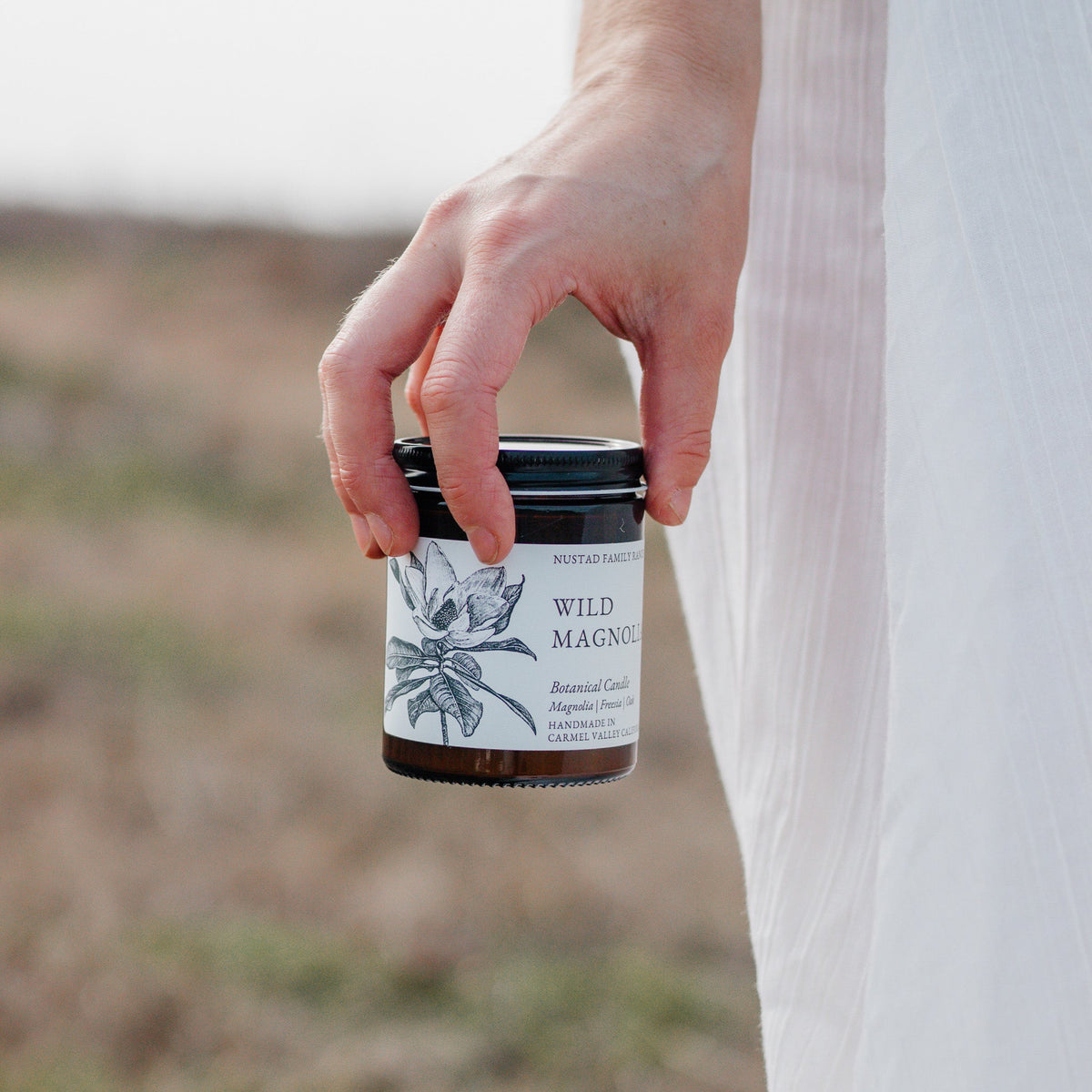 Person in a white dress holding an open Wild Magnolia botanical candle outdoors.