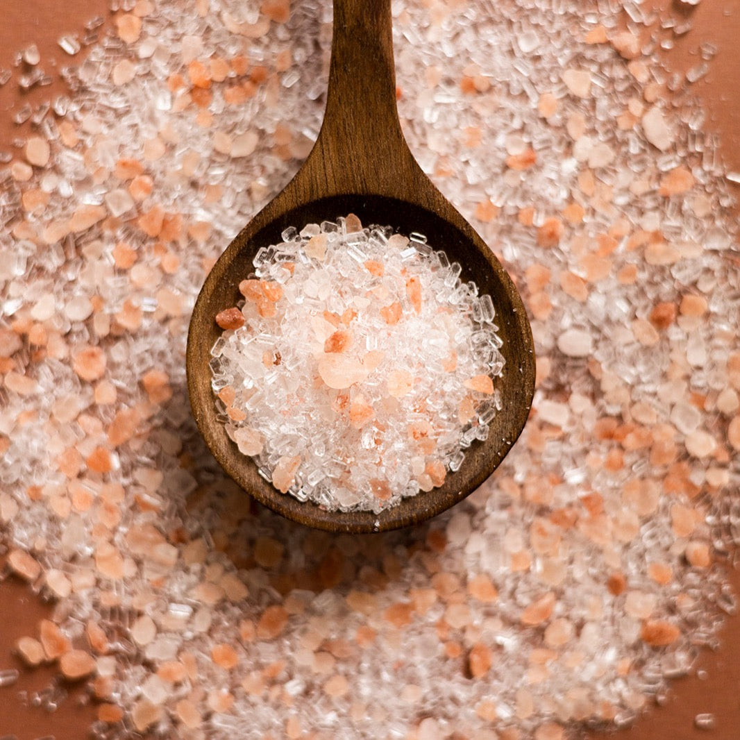 Wooden spoon filled with pink and white Himalayan salt crystals on a bed of salt. Kraft paper packaging of 'Afterglow' bath salts with a white label on a white background. essential oils epsom salt bath soak